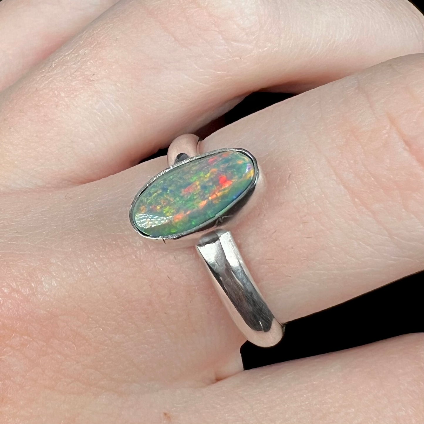 Angled close-up of a handmade sterling silver Australian dark opal solitaire ring with a red flash of color, worn on a finger.  Tooling marks are visible on the bezel.