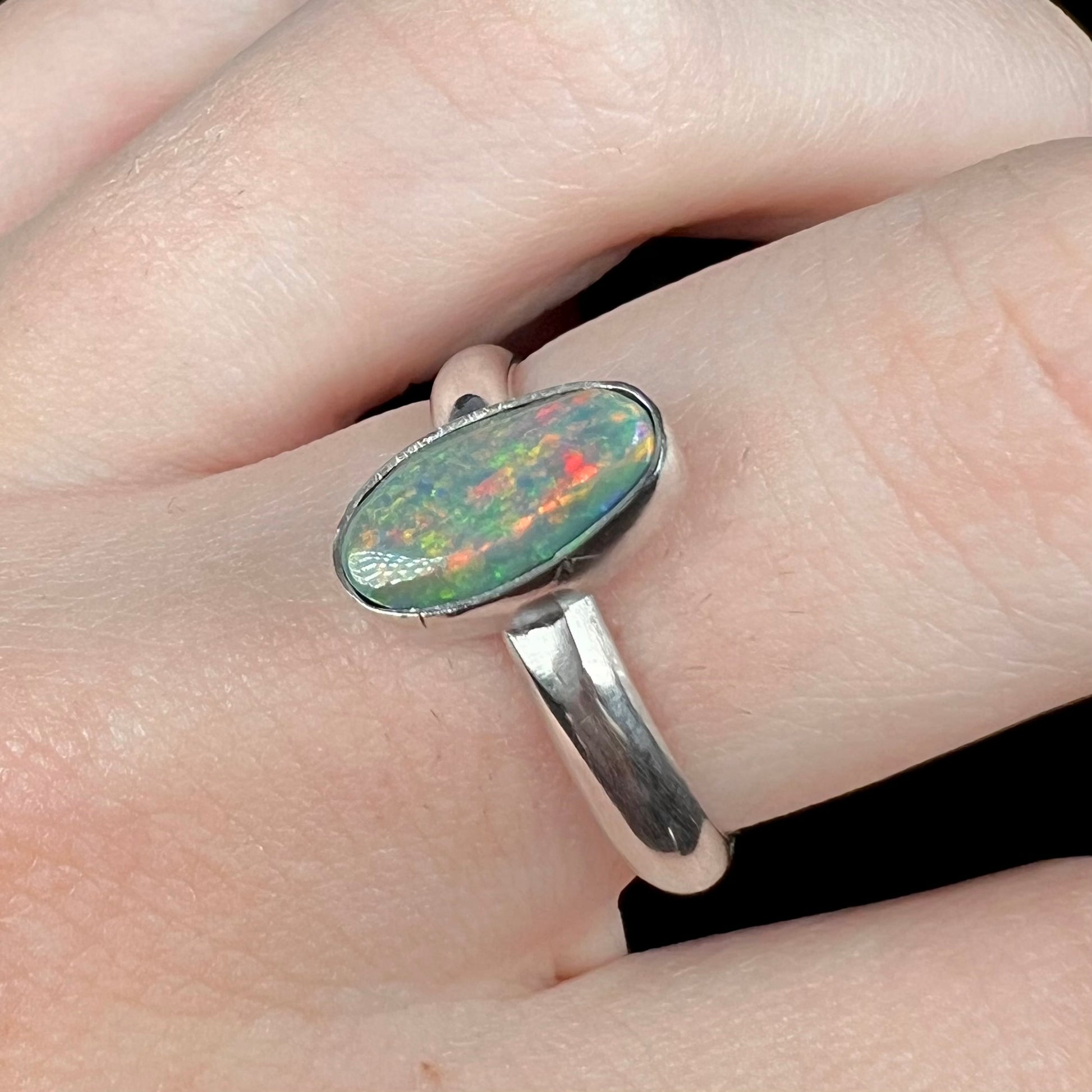 Angled close-up of a handmade sterling silver Australian dark opal solitaire ring with a red flash of color, worn on a finger.  Tooling marks are visible on the bezel.