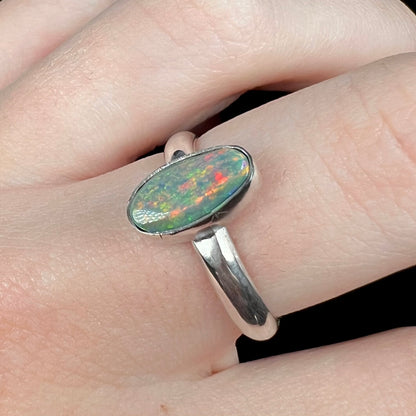 Angled close-up of a handmade sterling silver Australian dark opal solitaire ring with a red flash of color, worn on a finger.  Tooling marks are visible on the bezel.