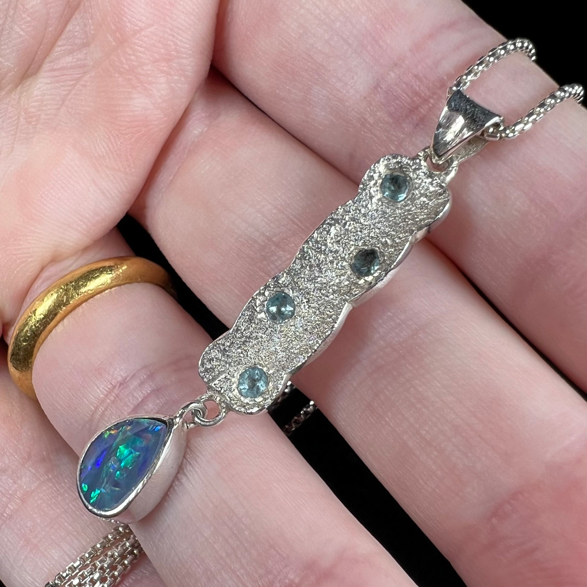 Angled close-up of a sterling silver Australian opal and blue topaz pendant on a chain, held between fingers under cool LED light.
