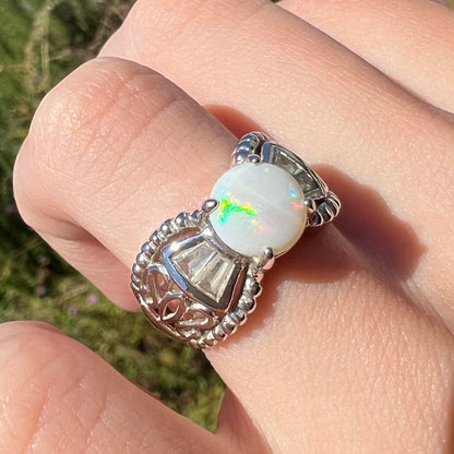 A round Australian white opal and CZ ring in white base metal worn on a finger, shown at an angle in the sun.