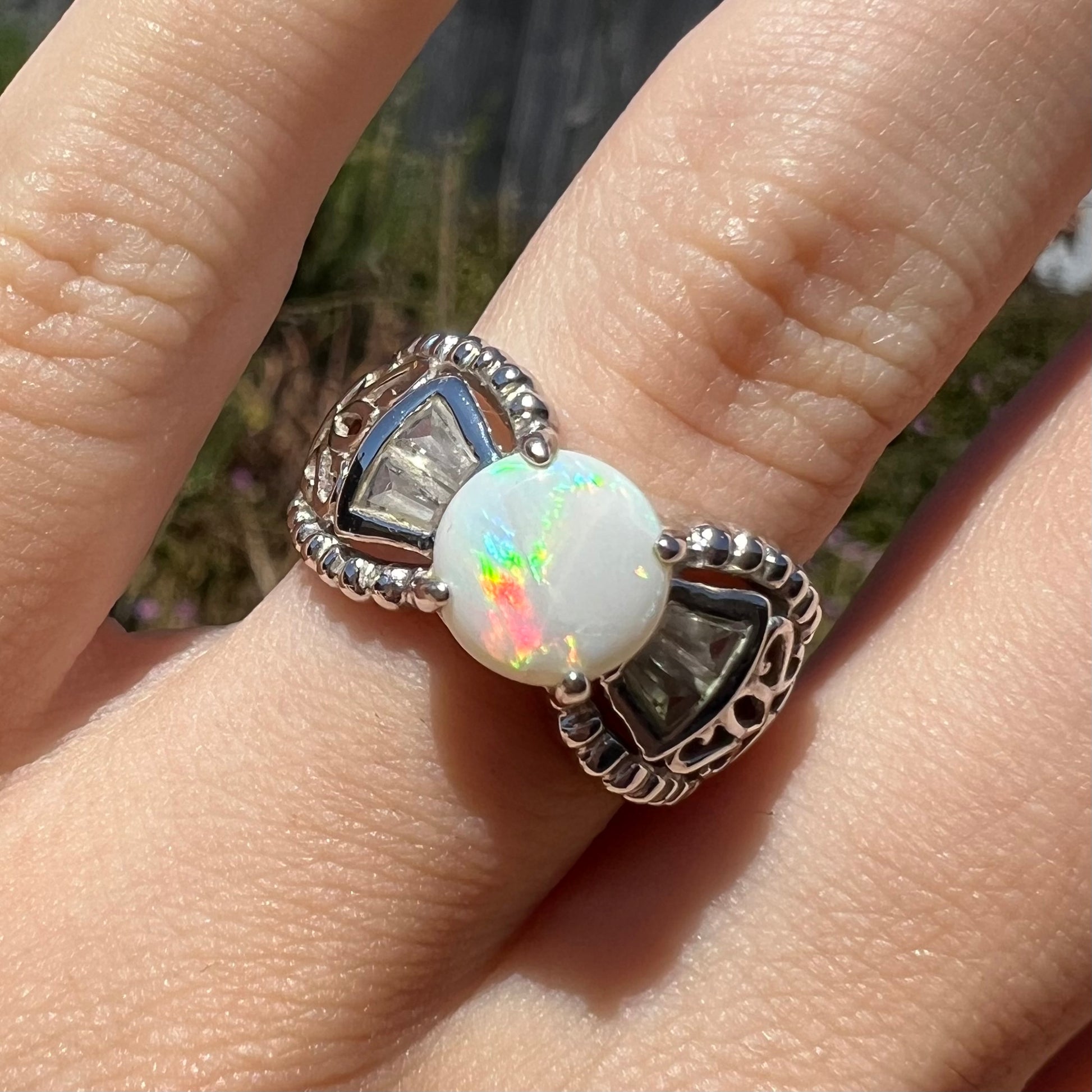 Close-up sunlit front view of a round Australian white opal and CZ ring in white base metal.  Opal has red flash.