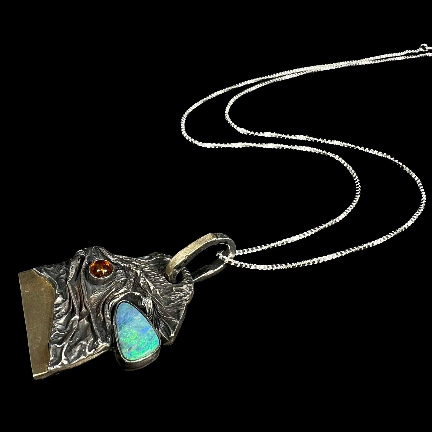 A brutalist-style Australian opal doublet and amber necklace in sterling silver and 18k gold laying on a black background, its chain flowing behind it.