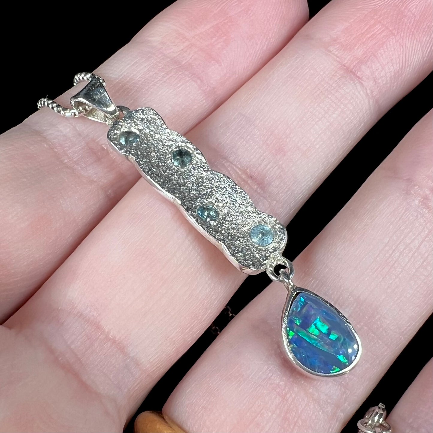 Teal flashes shine from an Australian opal doublet pendant in sterling silver with blue topaz accents and a textured design, resting on fingers.