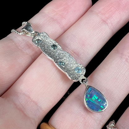 Teal flashes shine from an Australian opal doublet pendant in sterling silver with blue topaz accents and a textured design, resting on fingers.