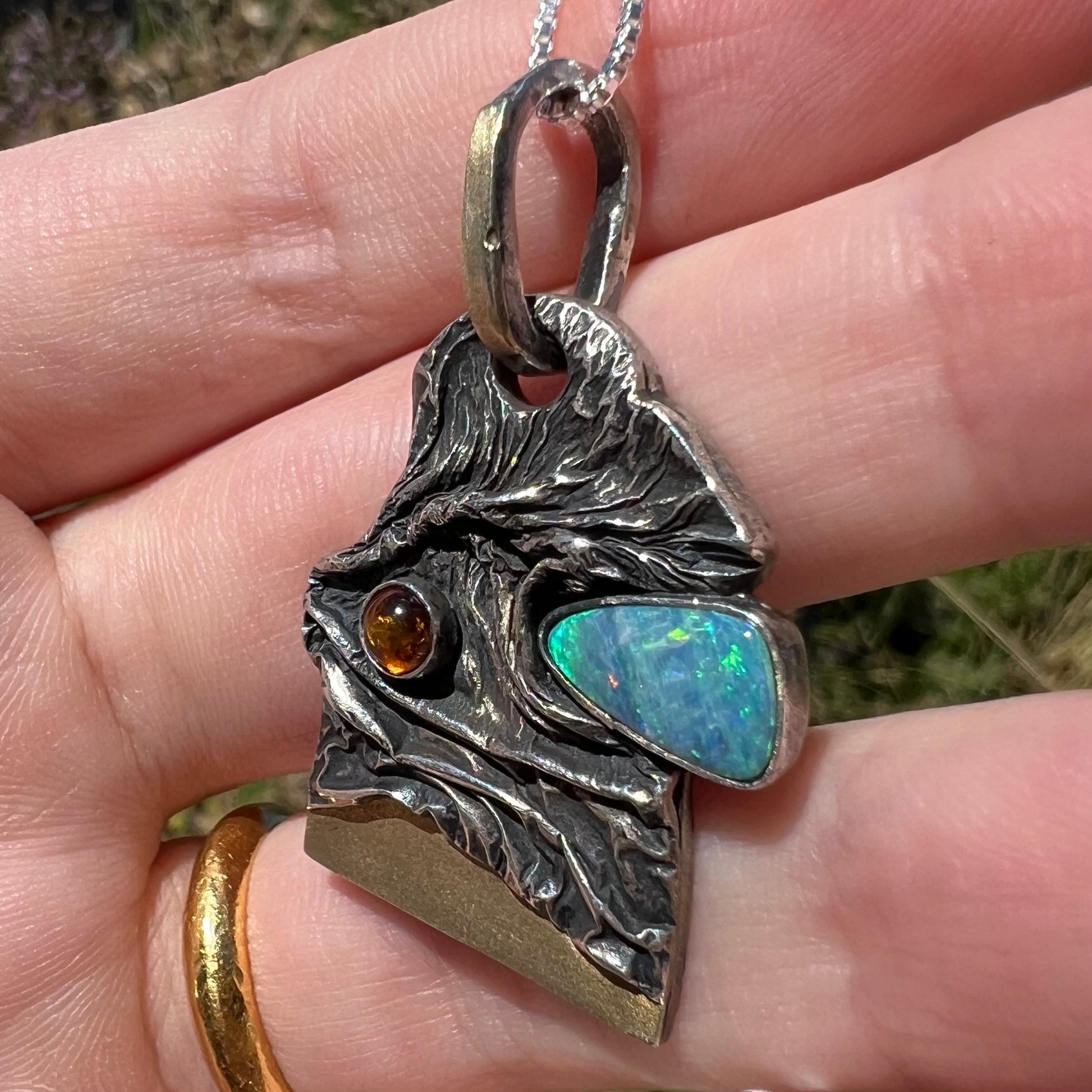 A rugged, rustic-style Australian opal doublet and amber men's pendant handmade from sterling silver with 18k gold accents, held from an angle in natural sunlight.