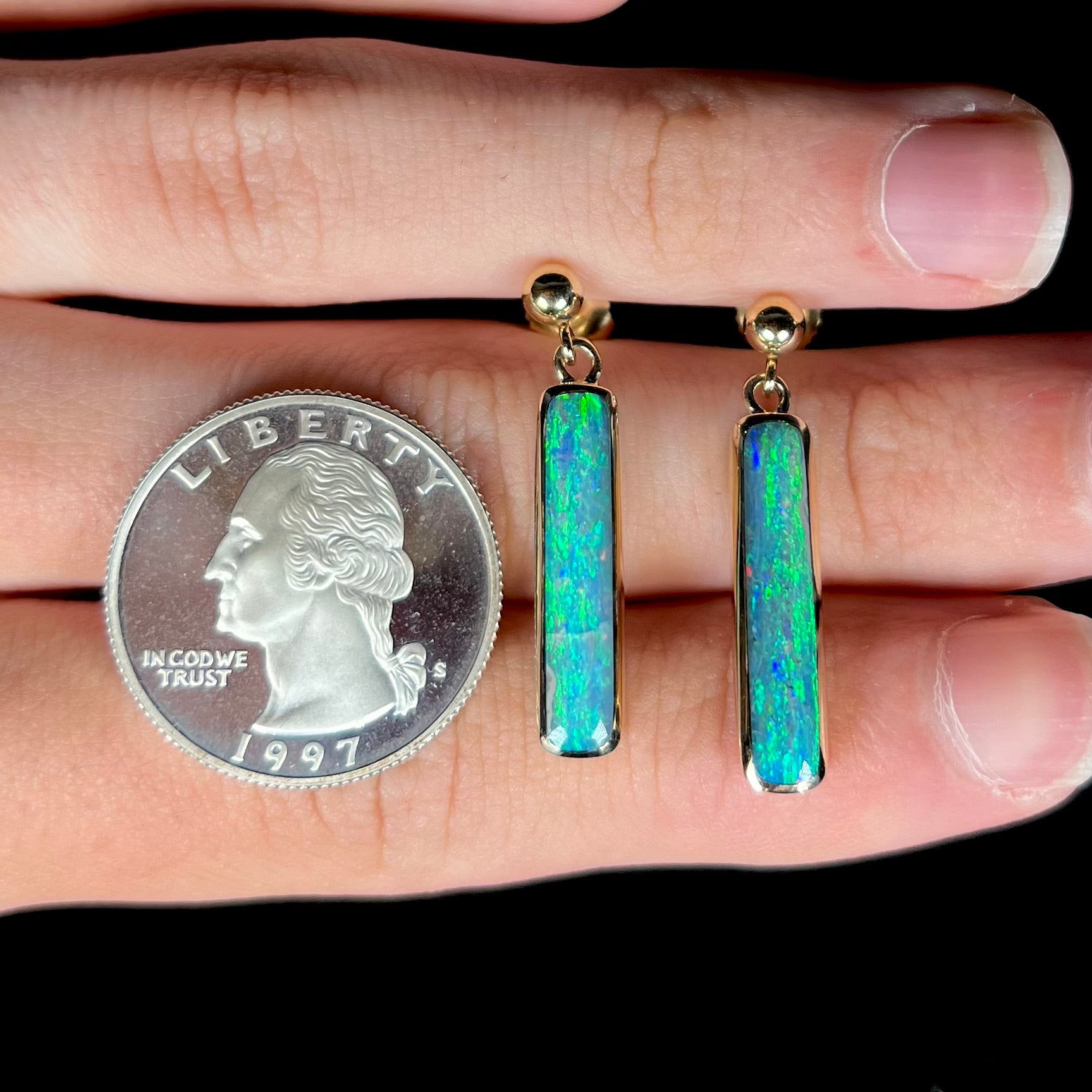 A pair of Australian opal drop earrings shown on top of a hand next to a 1997 S quarter coin for scale.
