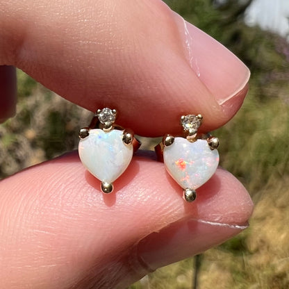 Close-up heart shaped Australian opal stud earrings with diamond accents in 14k yellow gold, shown held between two fingers in sunlight.