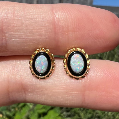 Close-up of Australian opal in onyx stud earrings held between two fingers in natural light against outdoor backdrop.