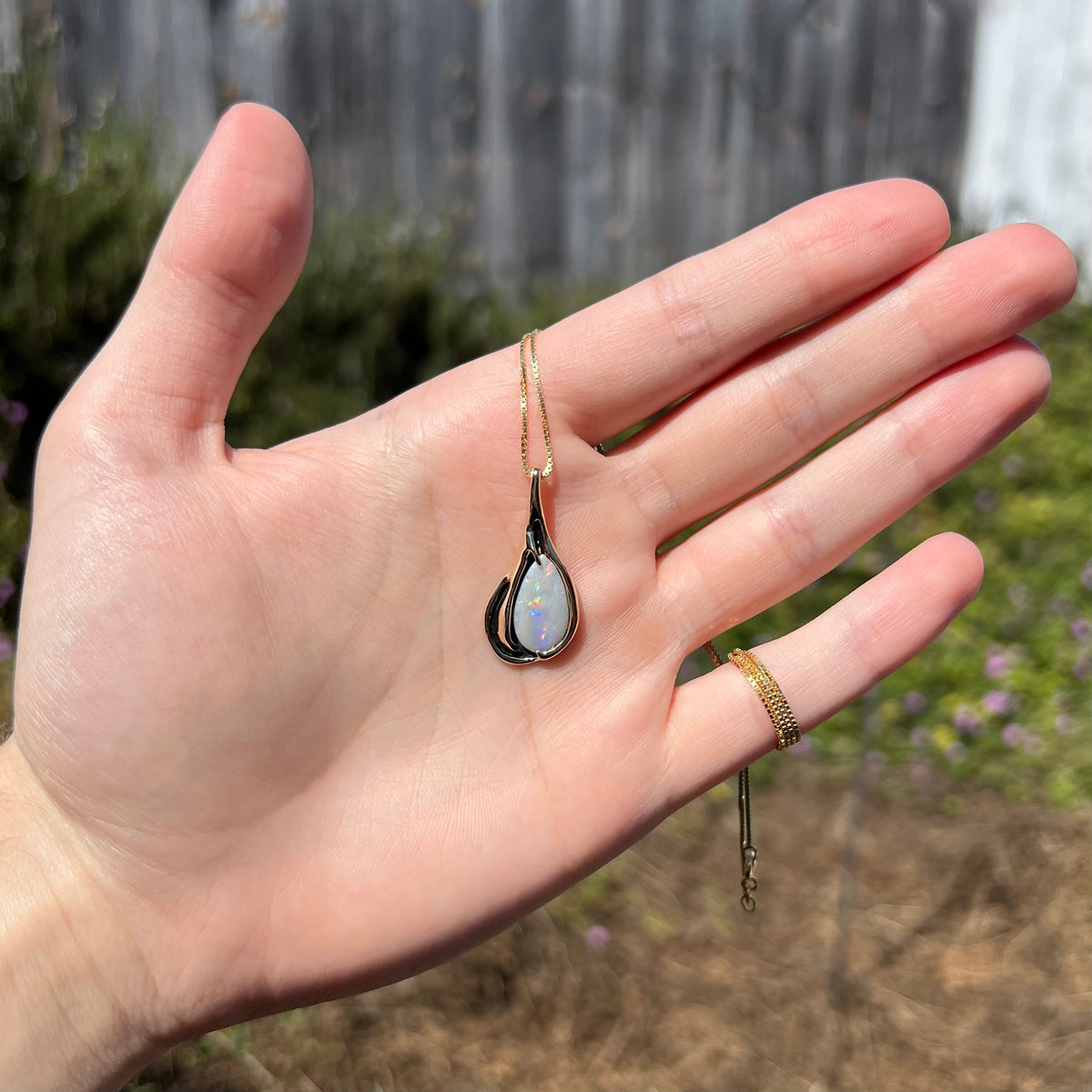 Sunlit outdoor view of pear shaped Australian opal pendant in 14k gold in Modernist setting with black antiquing, held in hand