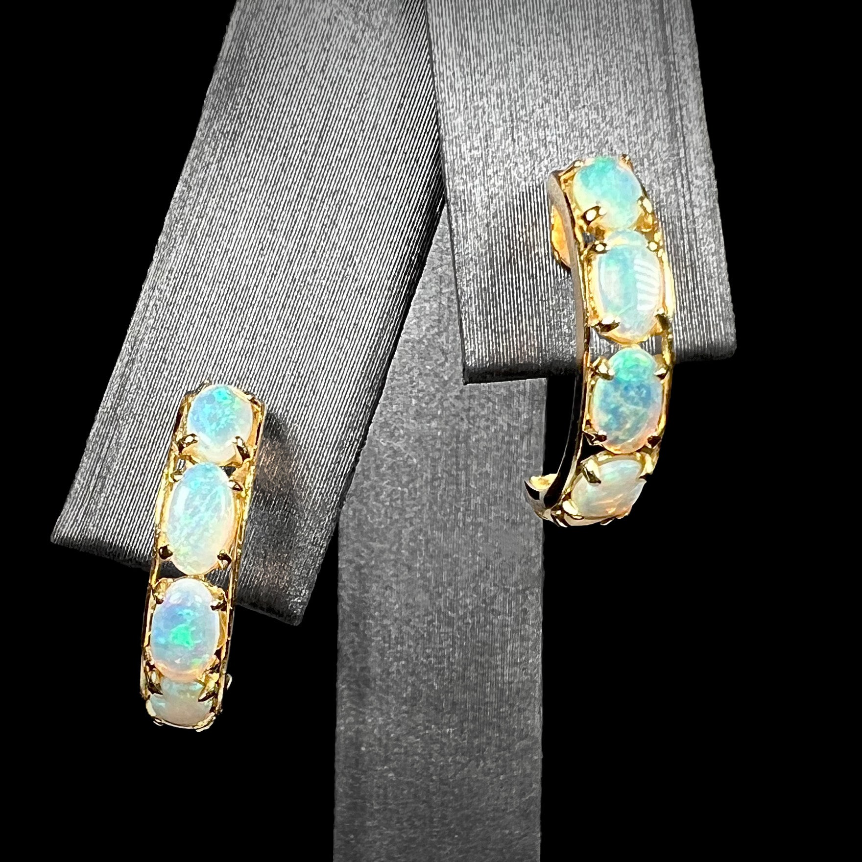 A pair of Australian crystal opal semi-hoop earrings in yellow gold shown on an earring display against a black background.
