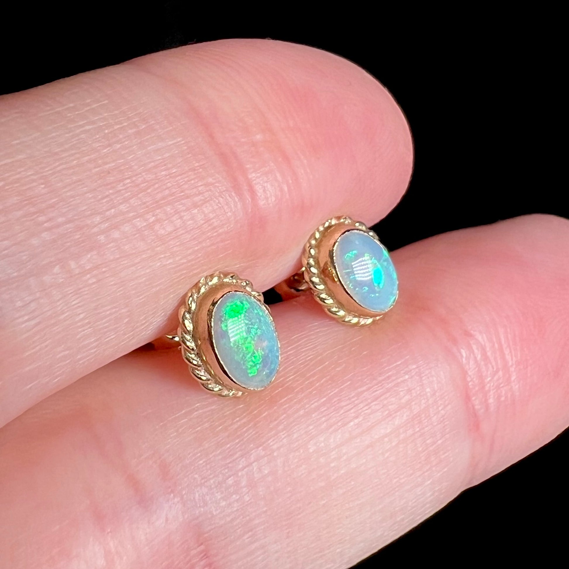 Angled close-up view of gold Australian opal stud earrings with rope bezel design, held between two fingers against black background.
