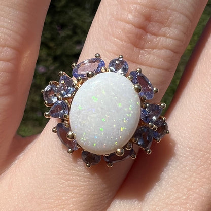 Close-up of Australian white opal ring with cluster of round, pear shaped, and oval tanzanite accents in 14k yellow gold prongs, worn on hand in sunlight