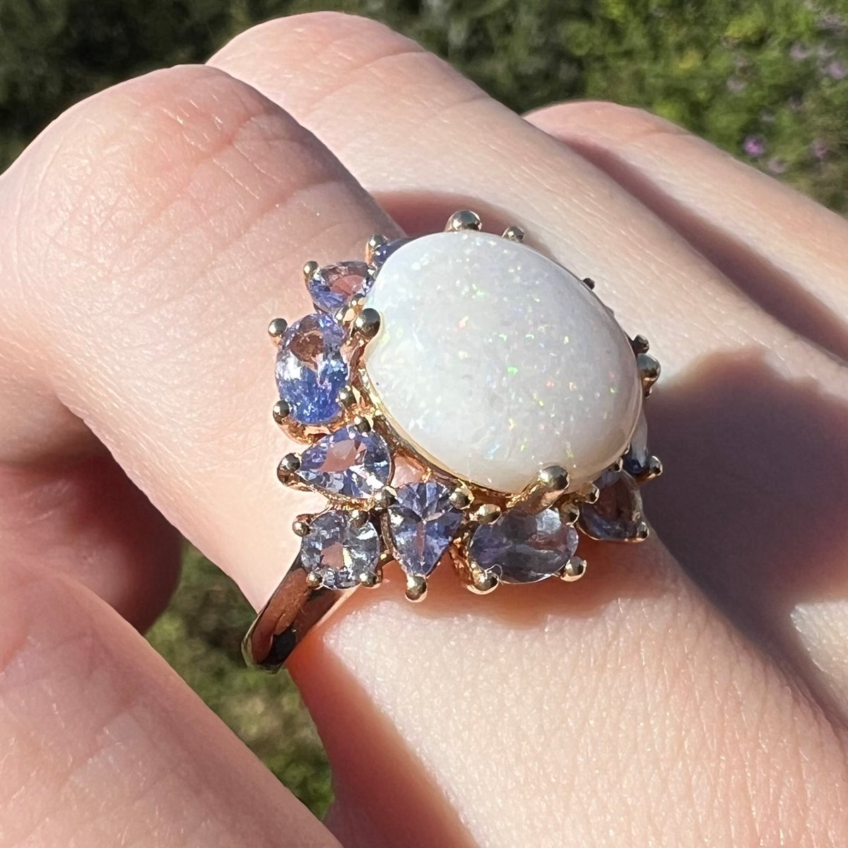 Angled macro view of Australian opal ring with cluster of different shaped violet tanzanite accents in 14k yellow gold, worn on finger in natural light