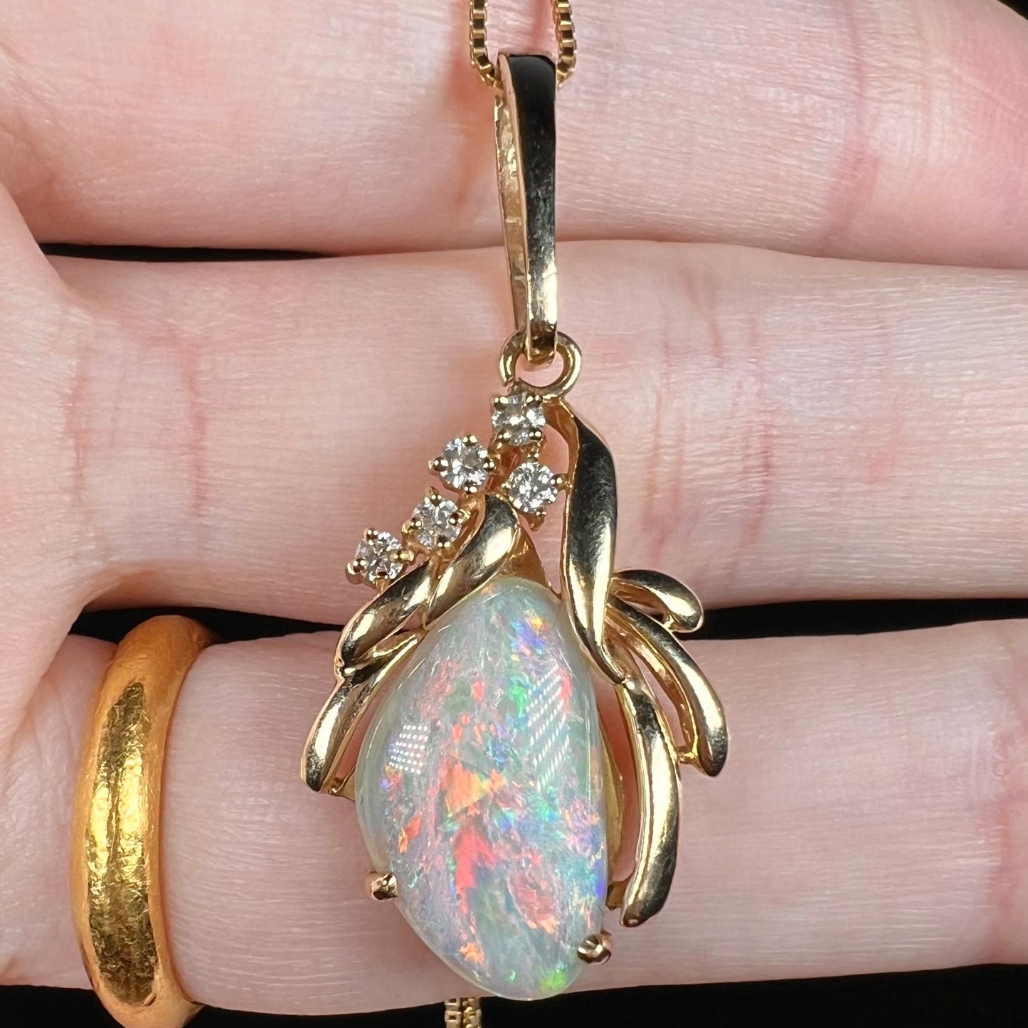 Multicolor red rainbow flashes are seen in an Australian shell opal pendant in 14k yellow gold with diamond accents, shown from a macro view in a hand.