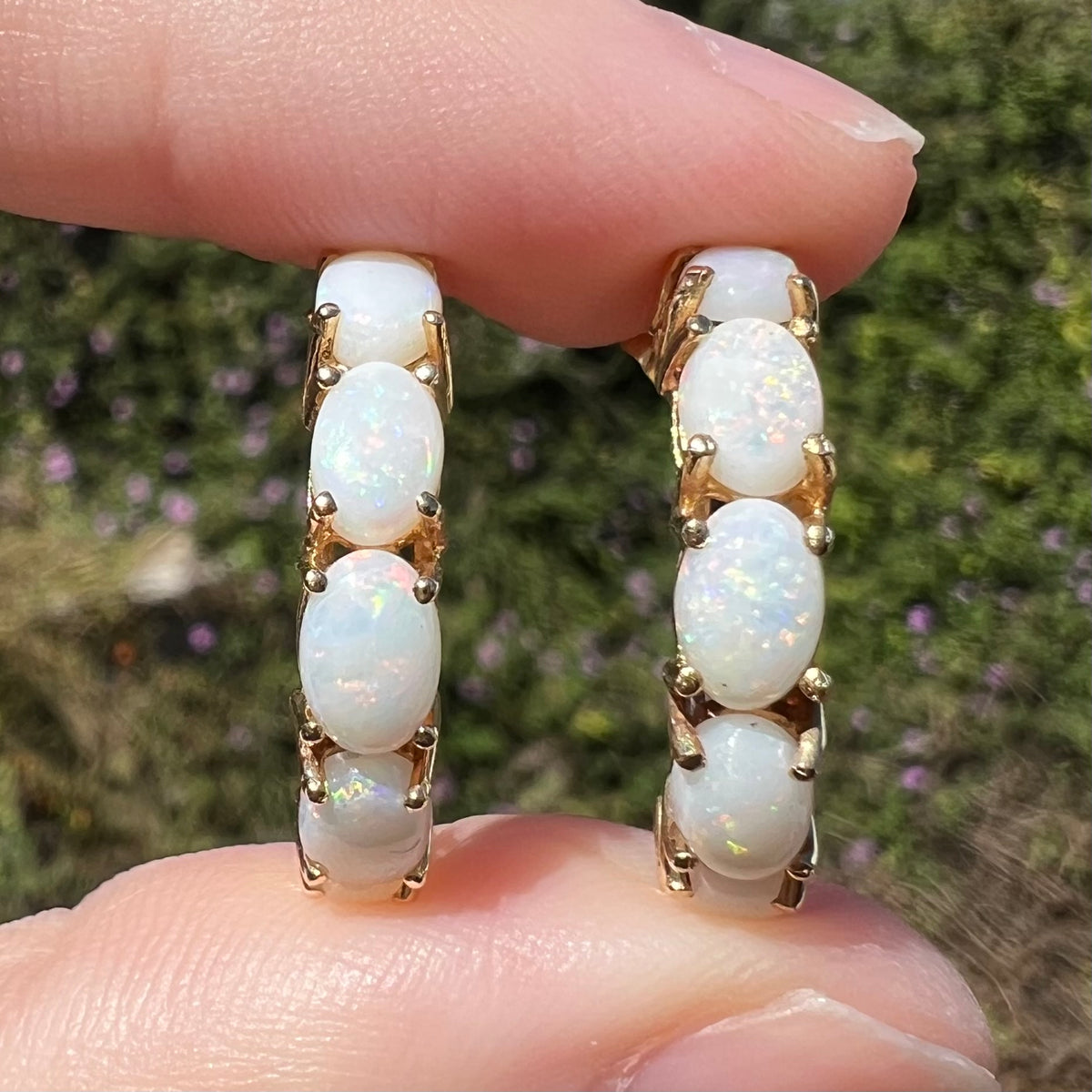 Close-up of women's Australian white opal semi-hoop earrings in yellow gold, held between two fingers in natural sunlight