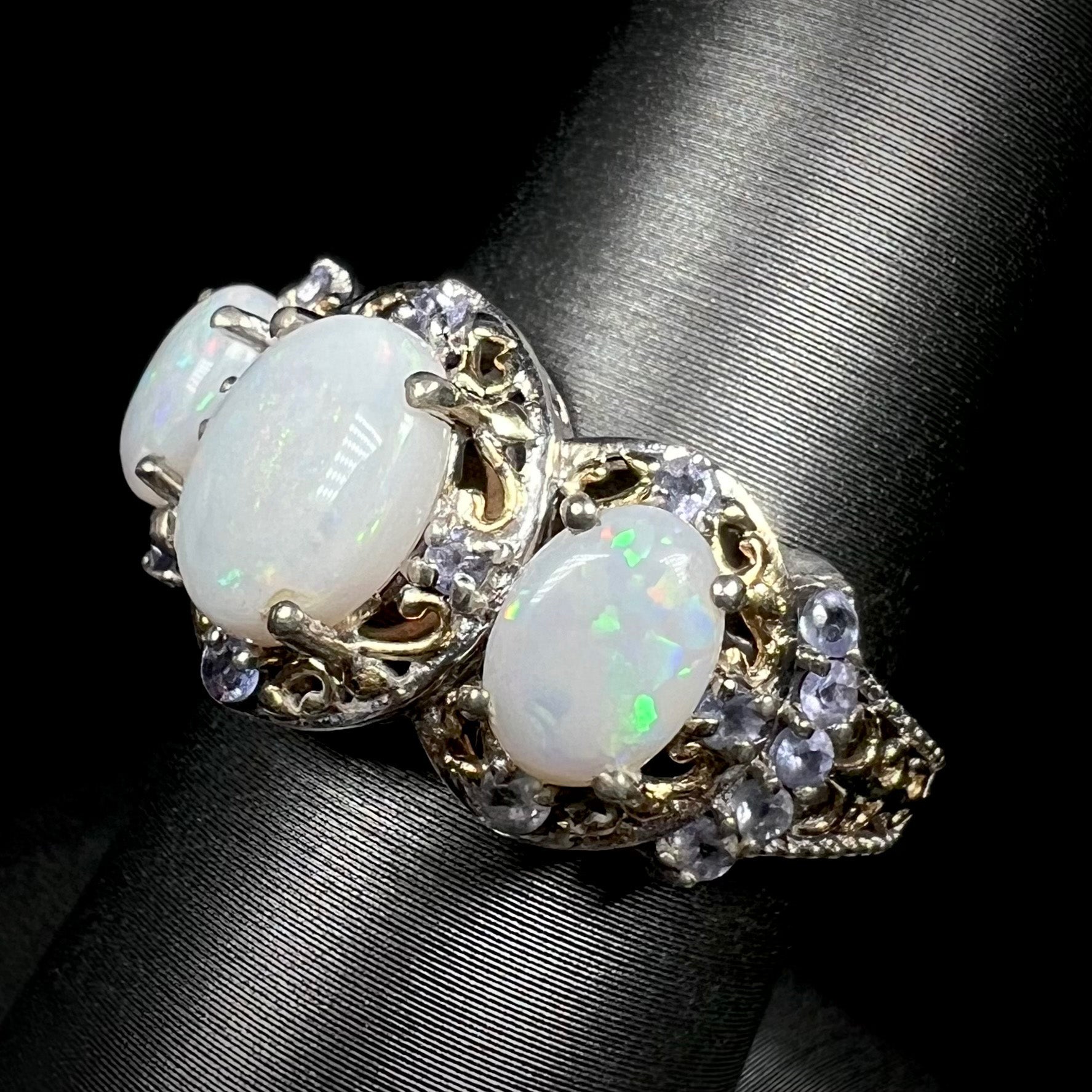 Angled close-up of an Australian white opal and tanzanite ring in sterling silver shown on ring display.  Yellow gold-plated two-tone filigree accents decorate the ring.