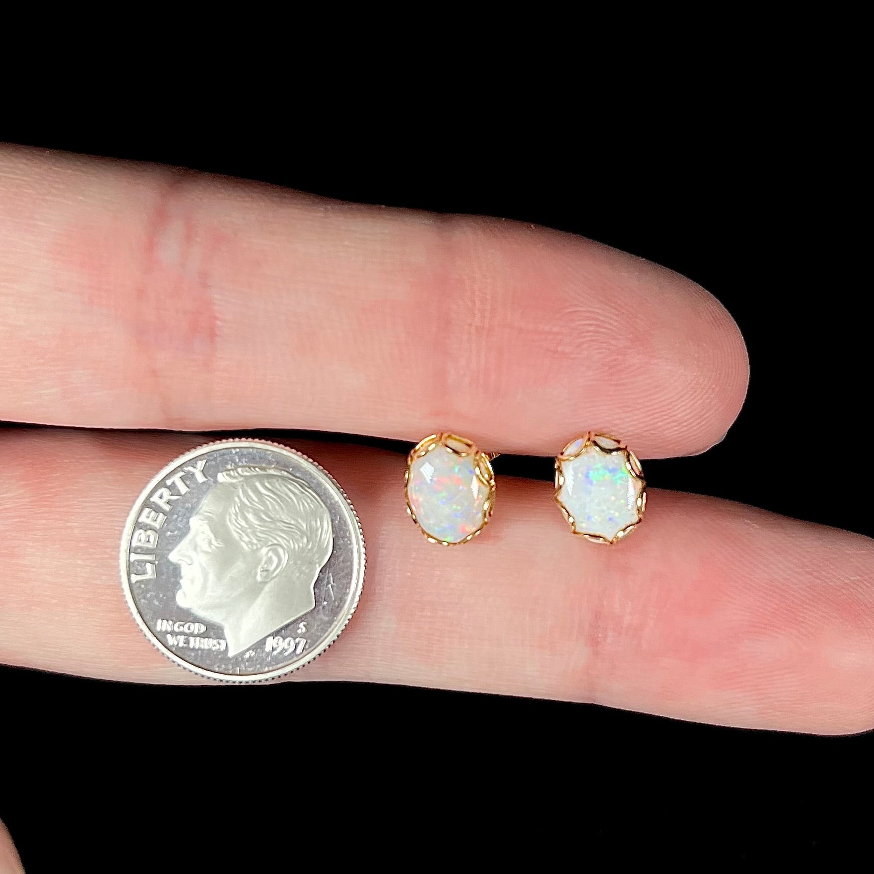 A pair of Australian white opal stud earrings in yellow gold shown between two fingers next to a dime coin.