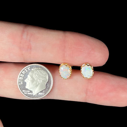 A pair of Australian white opal stud earrings in yellow gold shown between two fingers next to a dime coin.