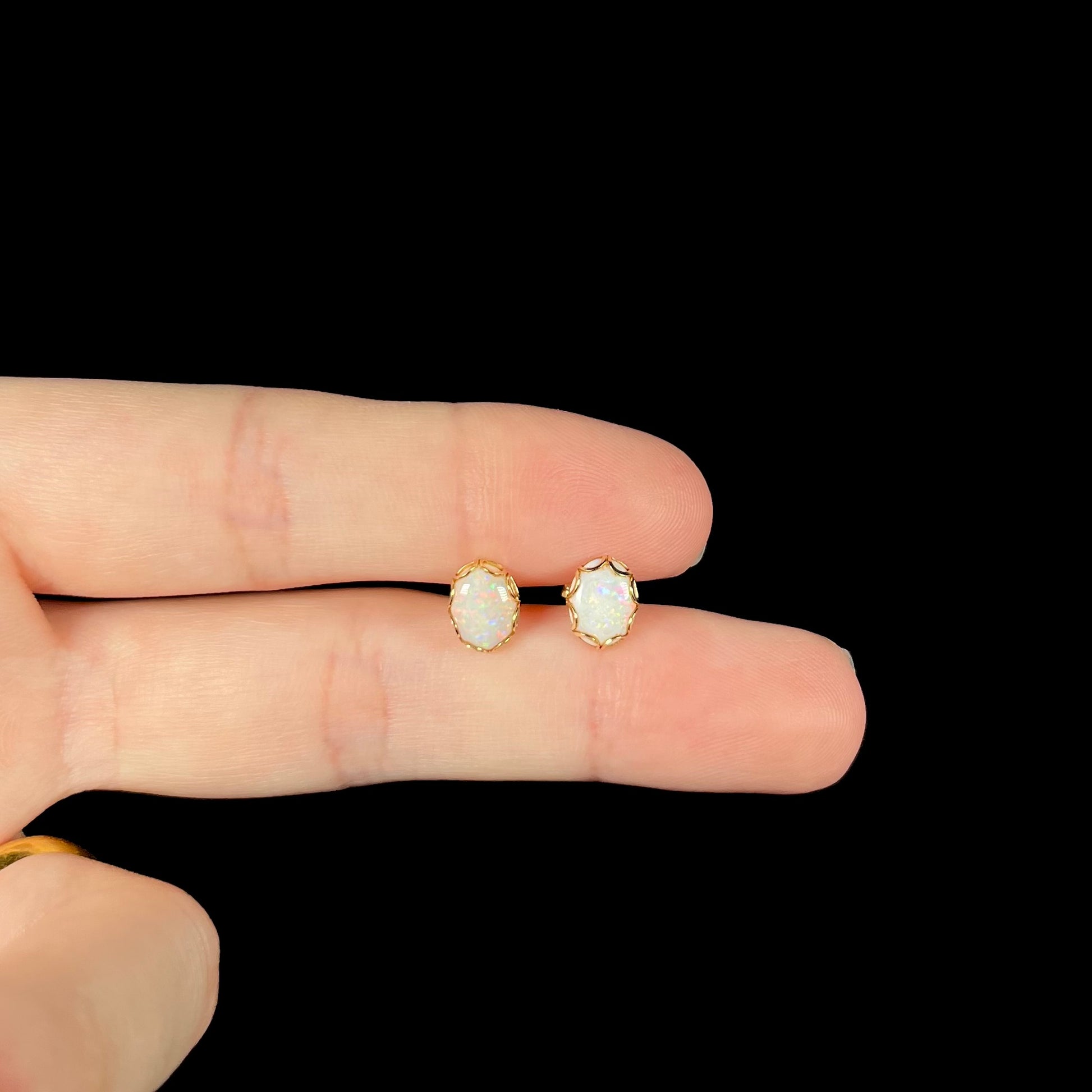 A pair of Australian white opal stud earrings in yellow gold shown held between two fingers against a black background.