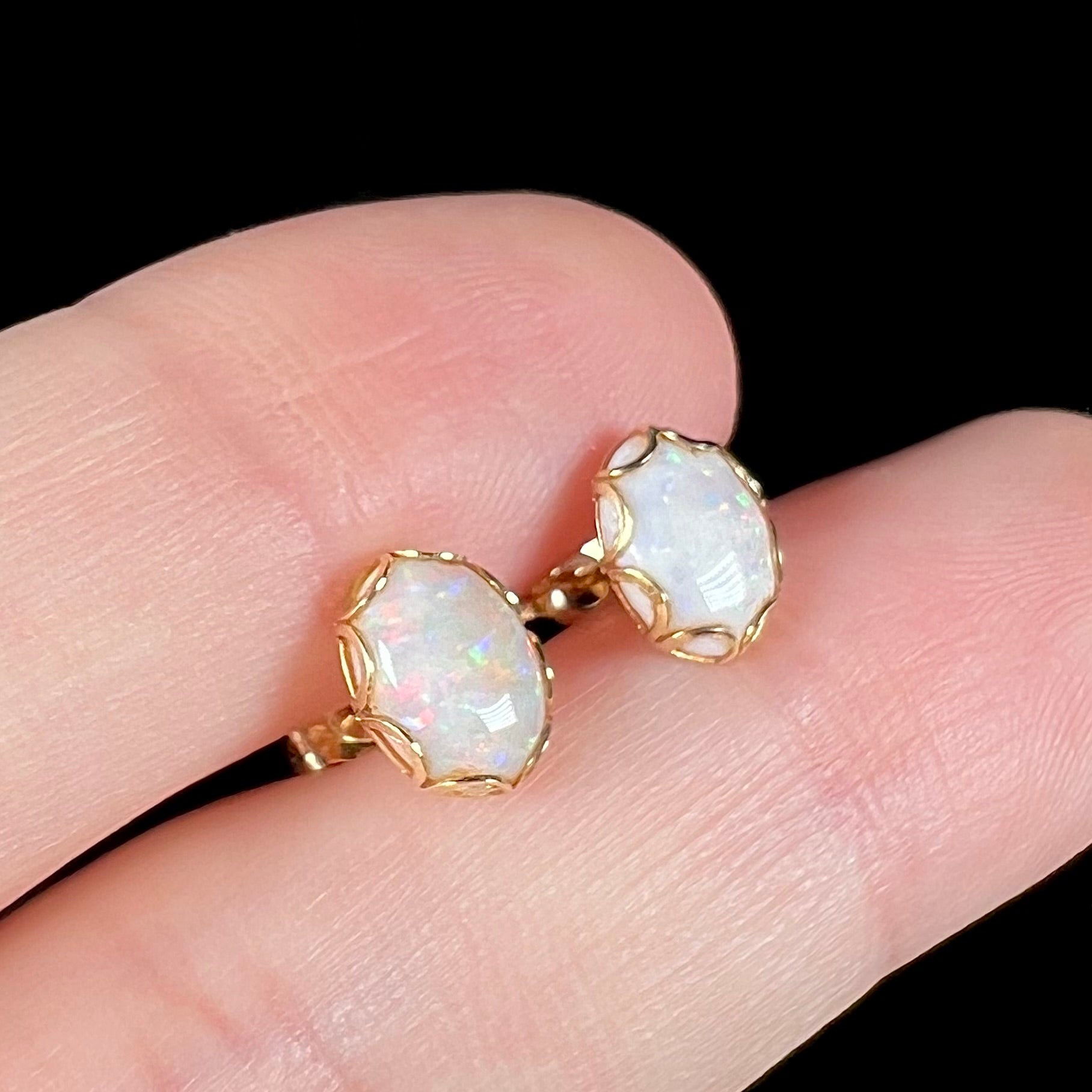 A pair of Australian white opal stud earrings in yellow gold shown close-up between two fingers from an angle.