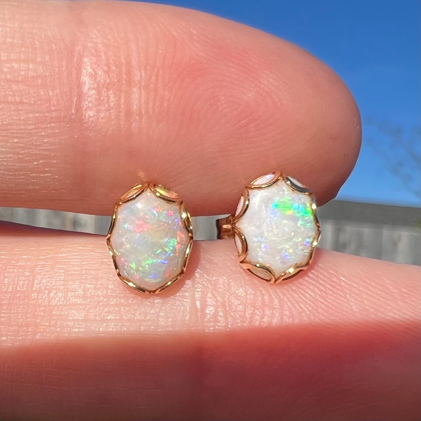 Close-up of a pair of Australian white opal stud earrings in yellow gold held between two fingers in sunlight.