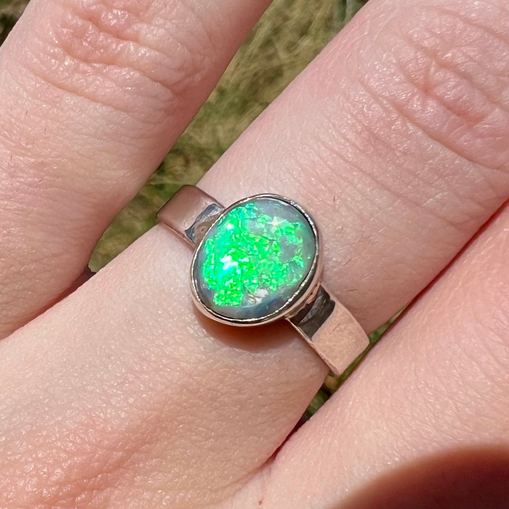 Close-up of a sterling silver Australian opal solitaire ring, worn on a finger in sunlight.  The opal has a bright, B4.5-rated flash of green play of color.