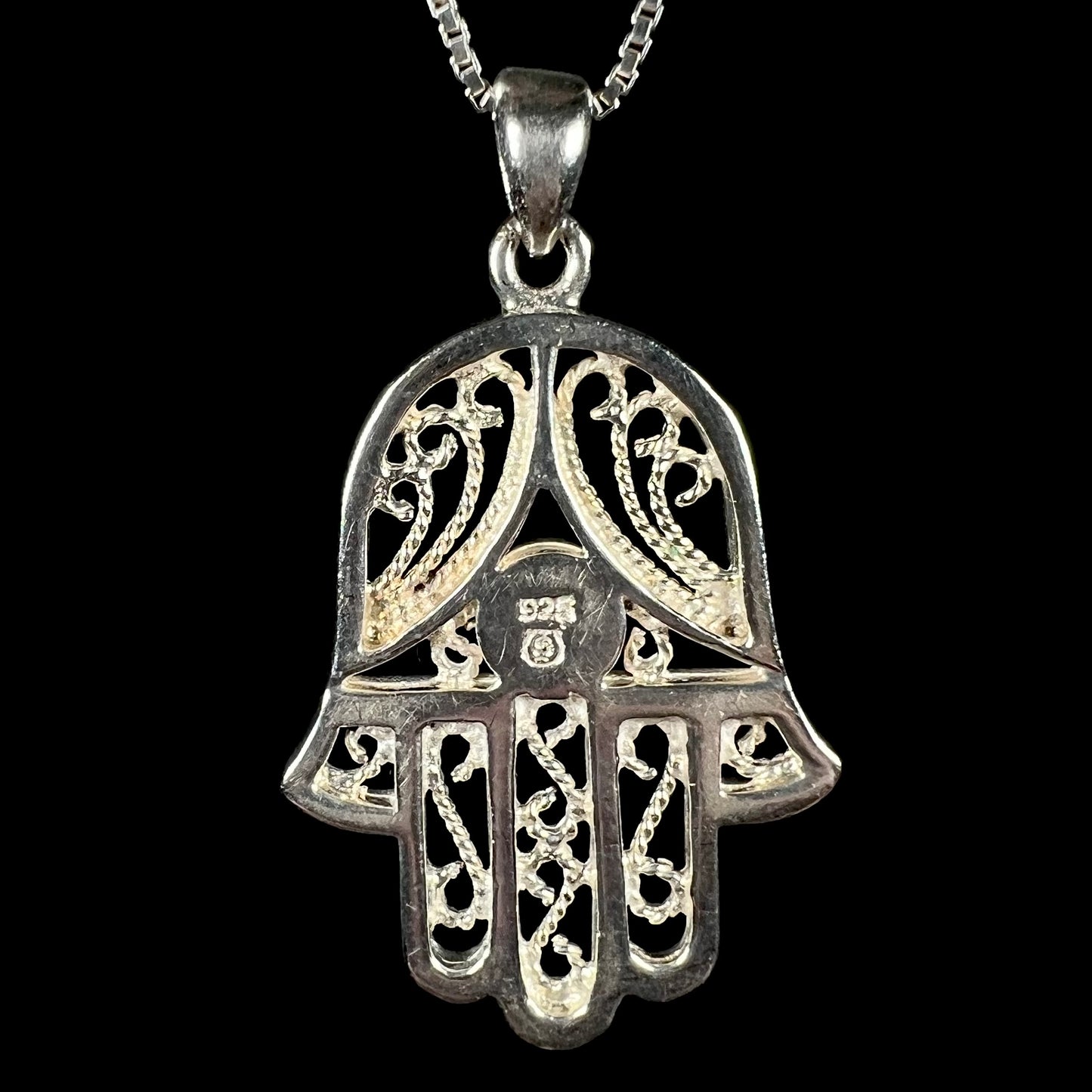Back of A sterling silver and filigree-accented hamsa hand necklace against a black background.