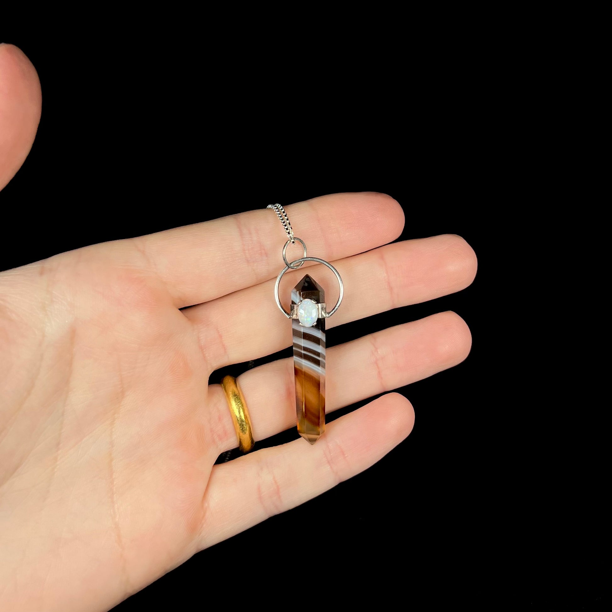 A banded sardonyx pendulum pendant with a white opal accent, held in a hand against a black background.
