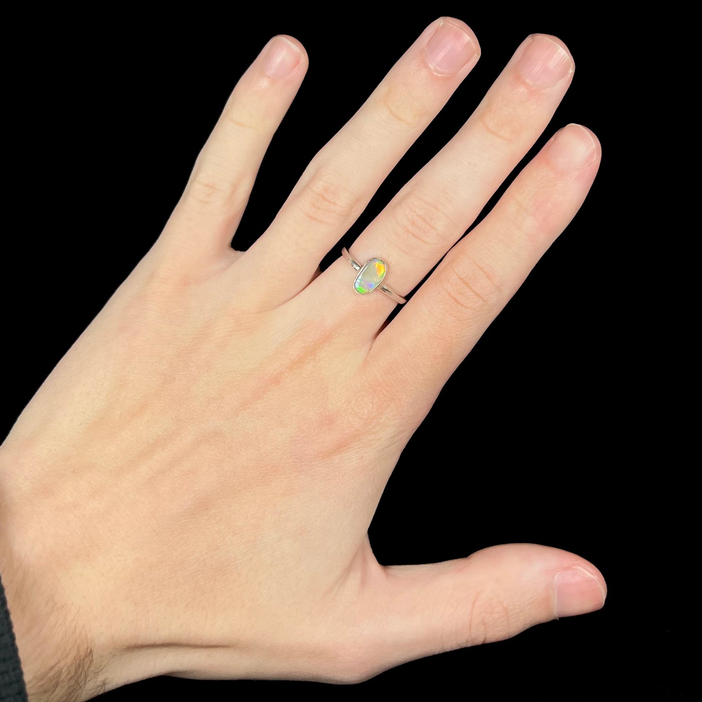 Hand wearing a bezel set sterling silver Australian opal solitaire women's ring against a black backdrop under cool LED lights.