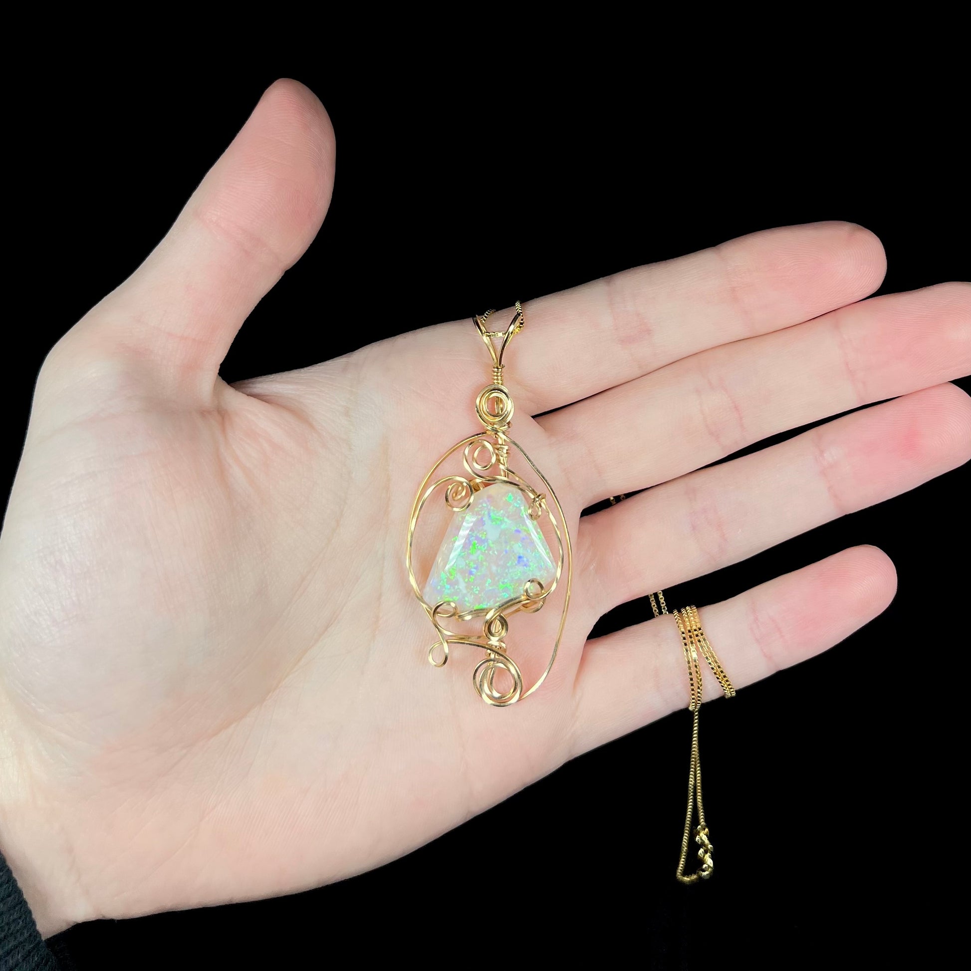 Hand holding a big Australian crystal opal pendant featuring a delicate gold-tone wire wrapped design beneath cool LED studio lights.