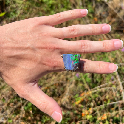 A big boulder opal ring with bright green fire, worn on a hand in the sunlight against a natural backdrop.