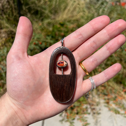 Hand holding a big men's fire opal and wood pendant in sterling silver in the sunlight.  The opal appears orange.