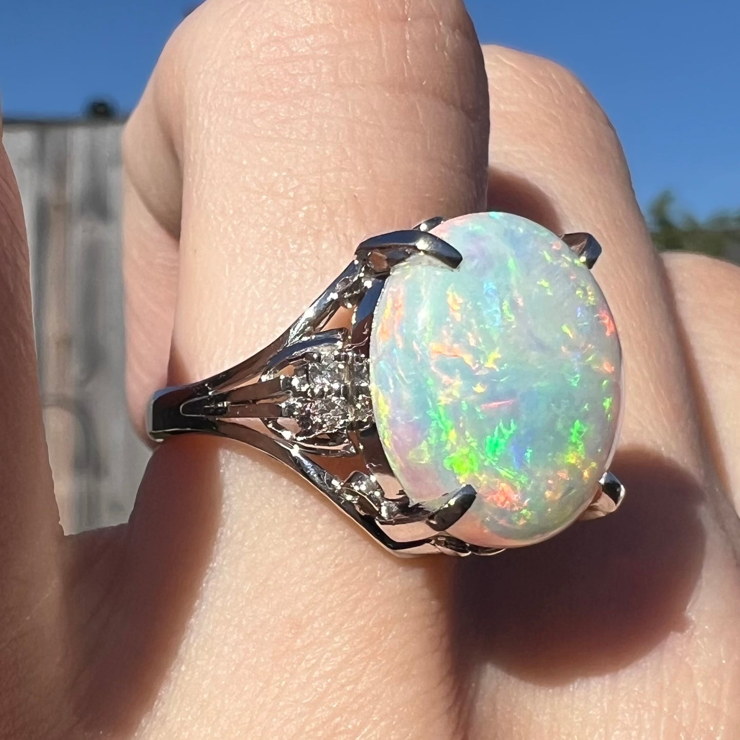 A big, 4.83ct platinum opal engagement ring with diamond accents worn on a finger in sunlight.