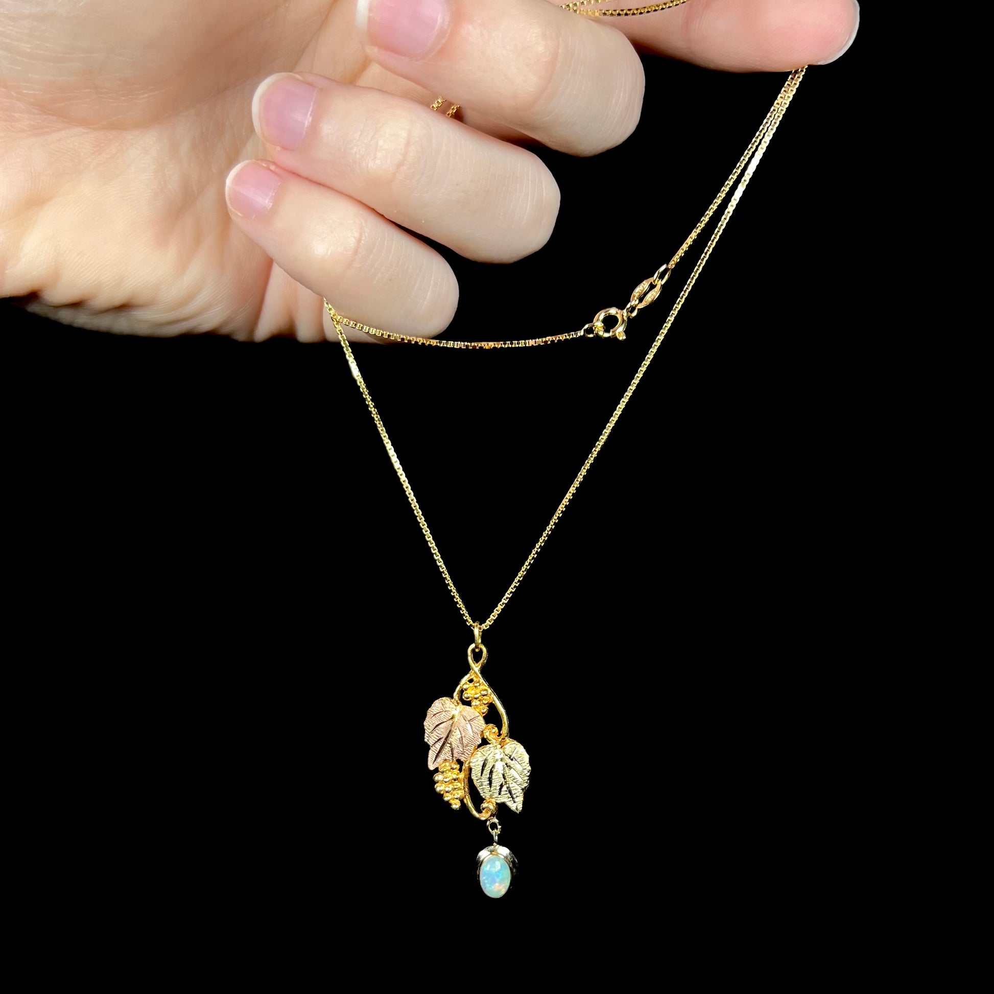 A Black Hills gold opal necklace being held by the chain against a black background, showing its detailed grapevine design.