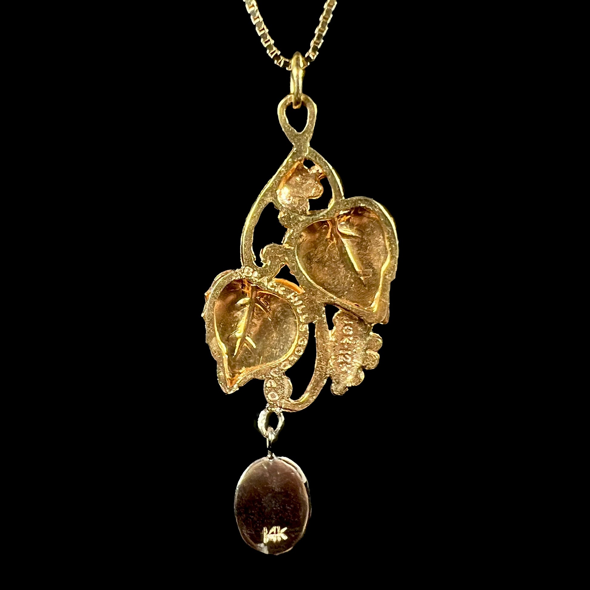 Back of a Black Hills gold pendant against a black background, displaying hallmark stamps “BLACK HILLS GOLD J CO”, “10K 12K”, and “14K”.