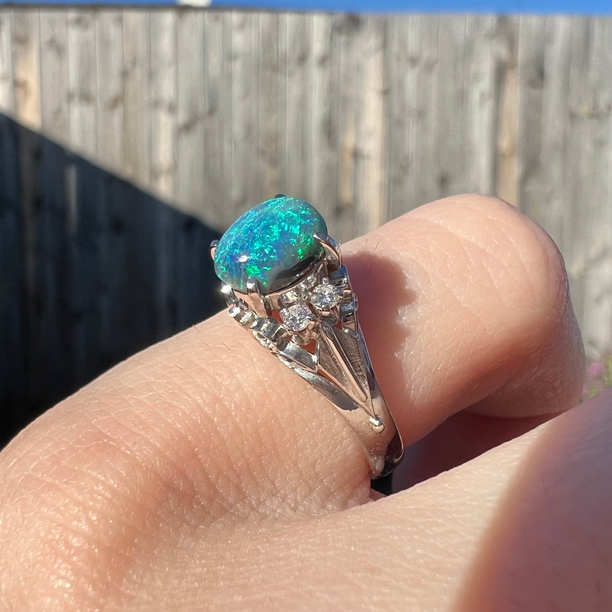 A black opal and diamond engagement ring worn on a finger in the sunlight.