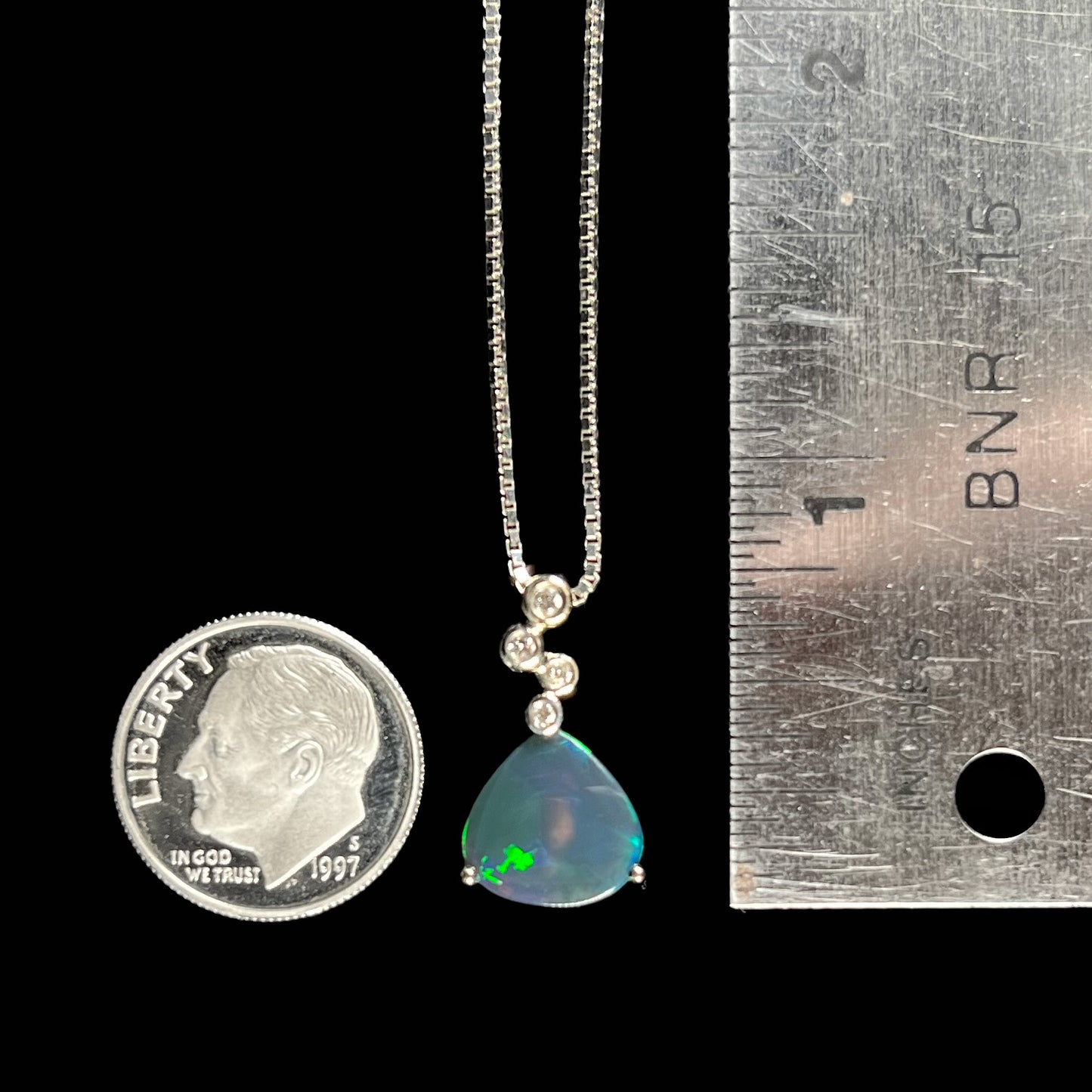 A black opal and diamond necklace next to a dime and ruler for size reference, measuring approximately 0.75 inches long.