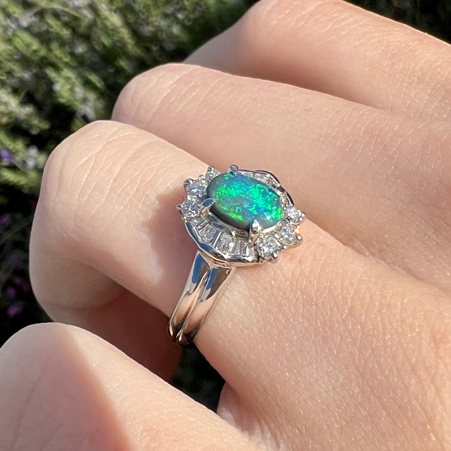 Blue-green fire is seen in a platinum black opal engagement ring, worn on a hand in the sun.  In-focus is the diamond baguette halo.