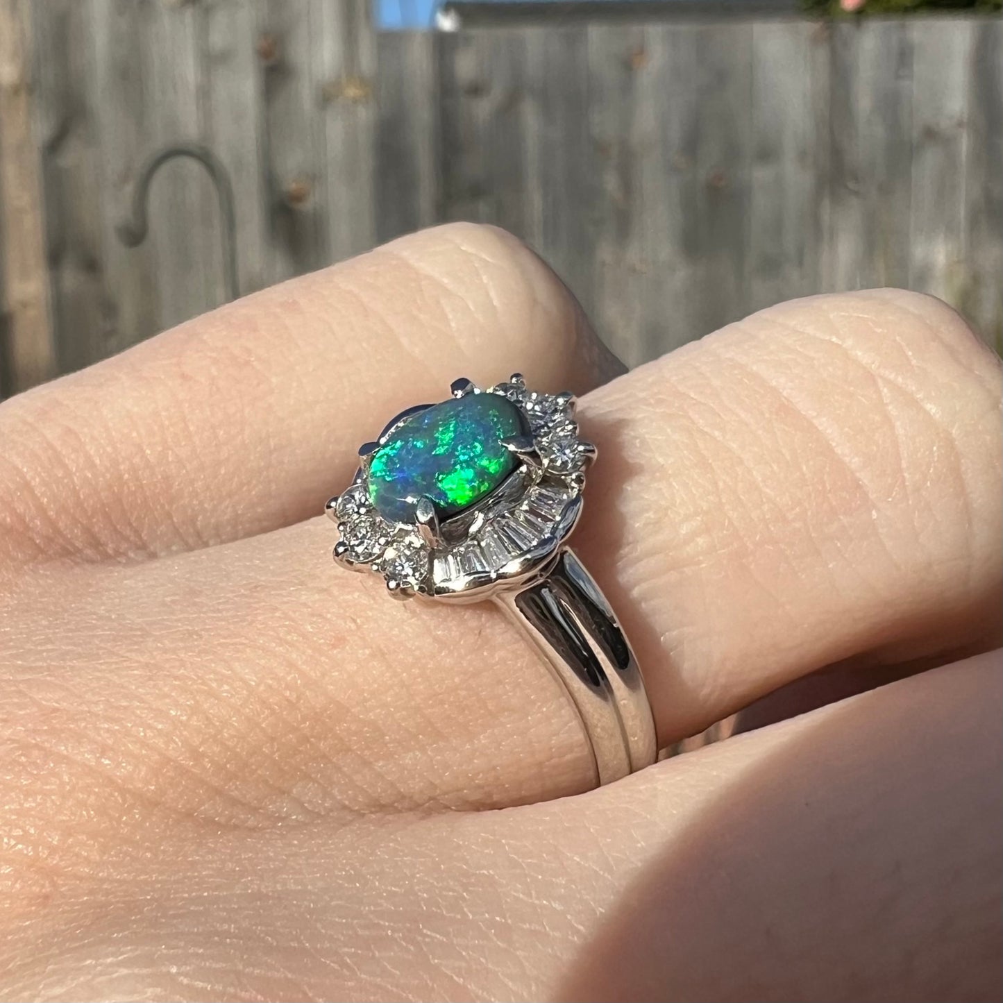 Angled view of a platinum black opal engagement ring, worn on a hand in sunlight.  Showcased are the baguette diamond halo and opal's green fire.