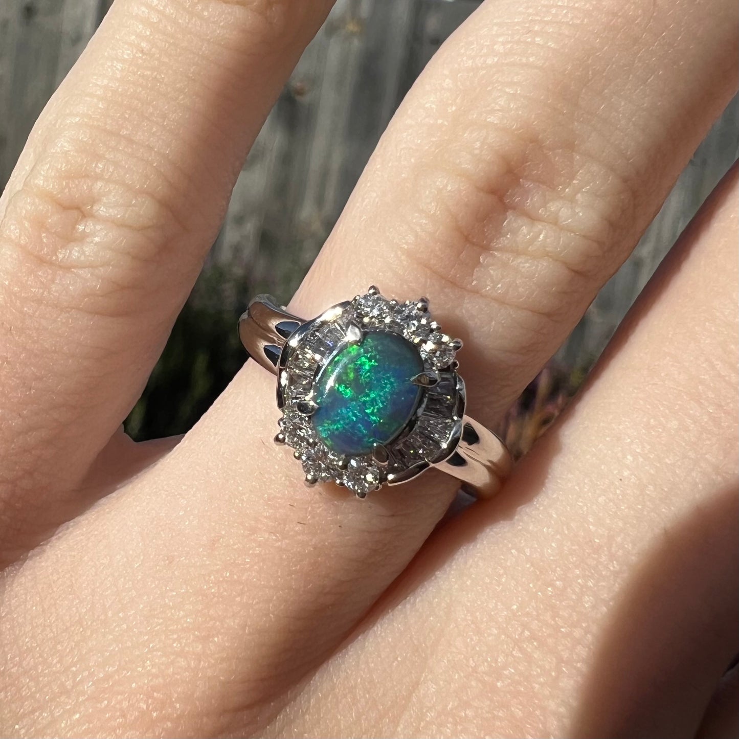 Close-up view of a platinum black opal engagement ring with a baguette diamond halo, worn on a finger in the sunlight.