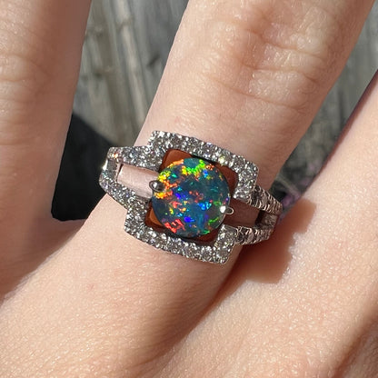 A Lightning Ridge black opal engagement ring with vivid red multicolor fire, set in a diamond-accented white gold ring.  Photographed in natural light on a hand for scale.