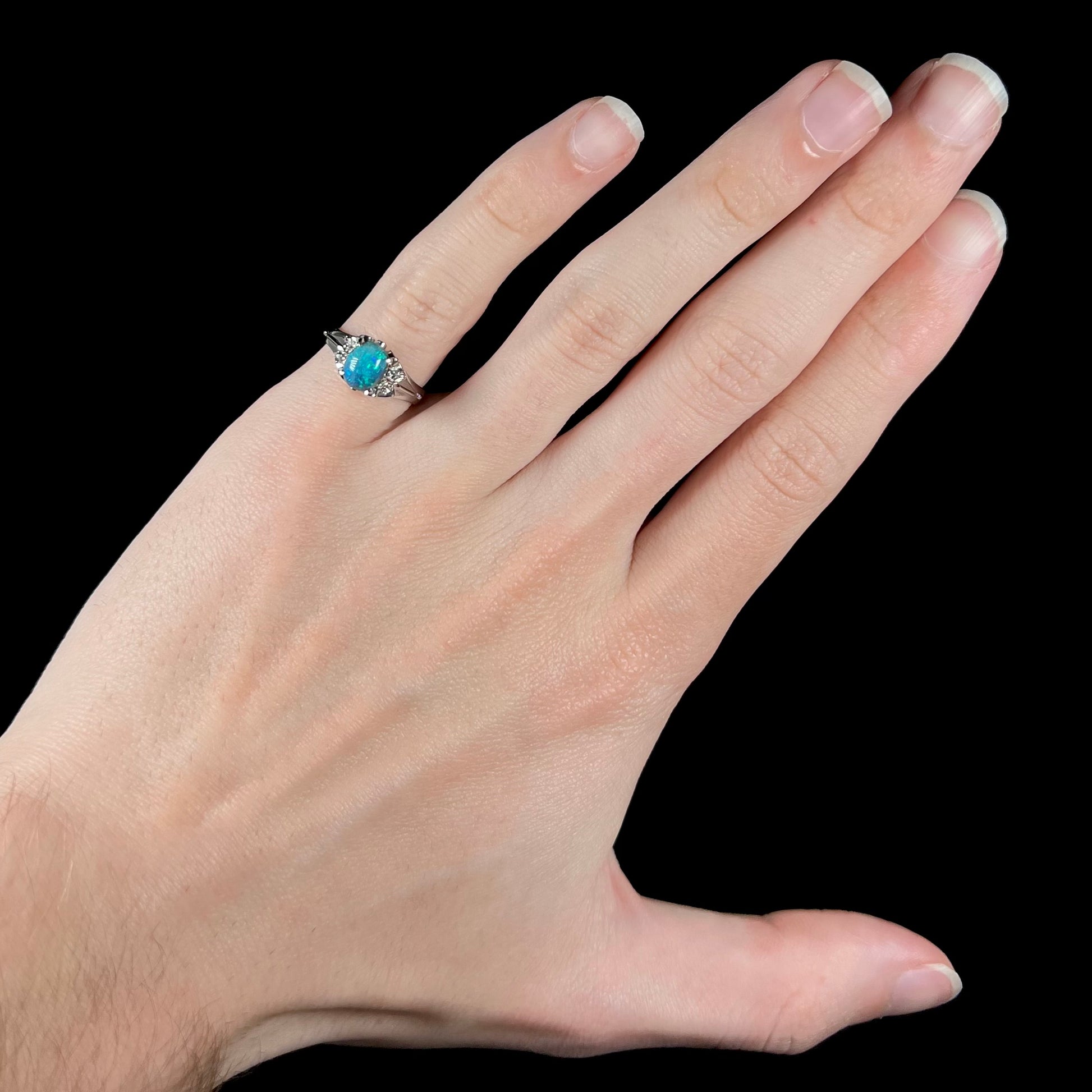 A dainty platinum black opal engagement ring with diamonds, worn on the pinkie finger of a hand.