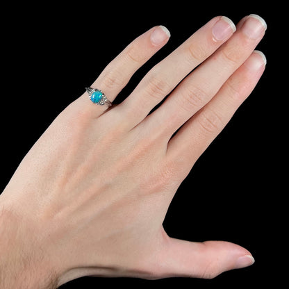 A dainty platinum black opal engagement ring with diamonds, worn on the pinkie finger of a hand.