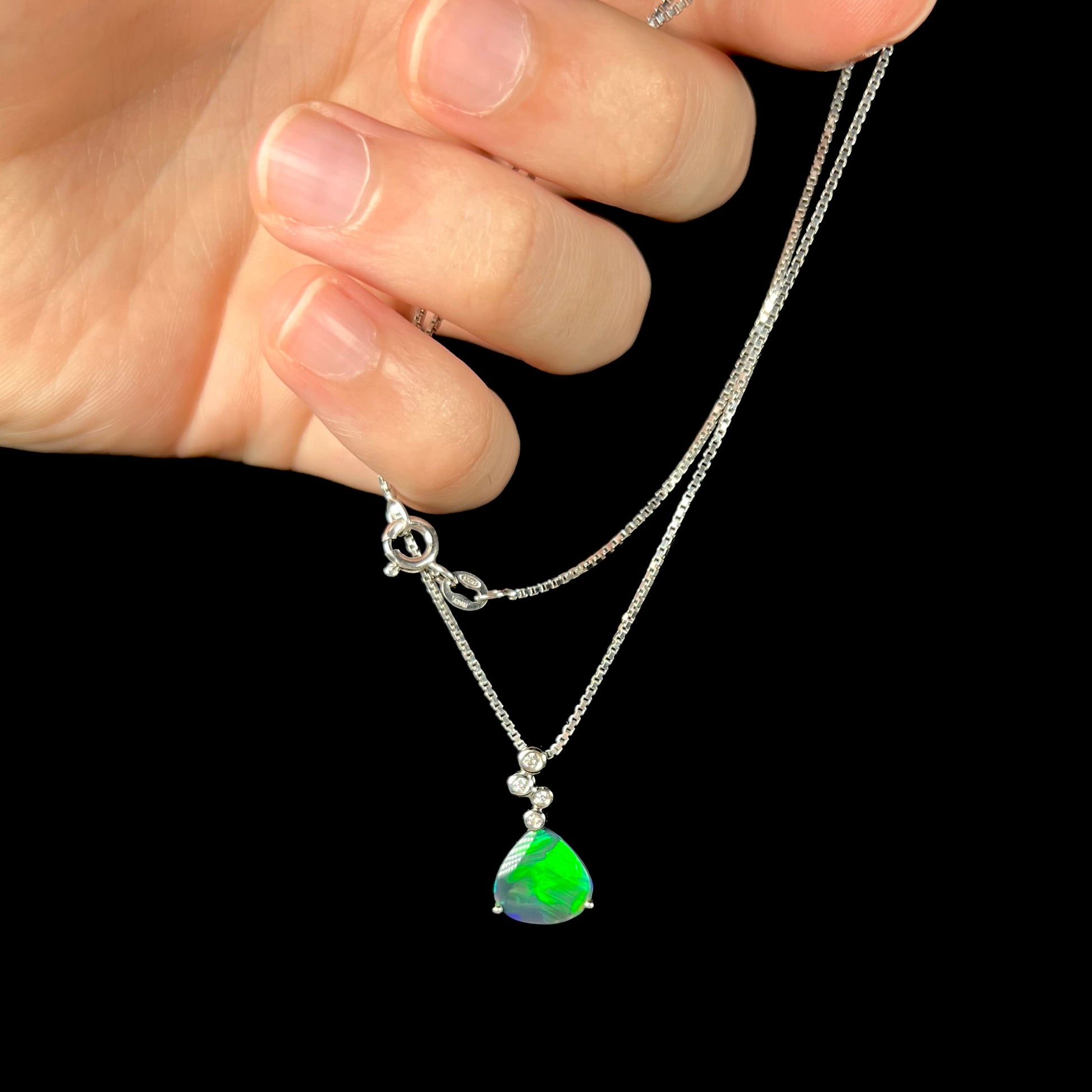 A black opal and diamond necklace being dangled in a hand against a black background, with a vibrant green flash visible in the opal.