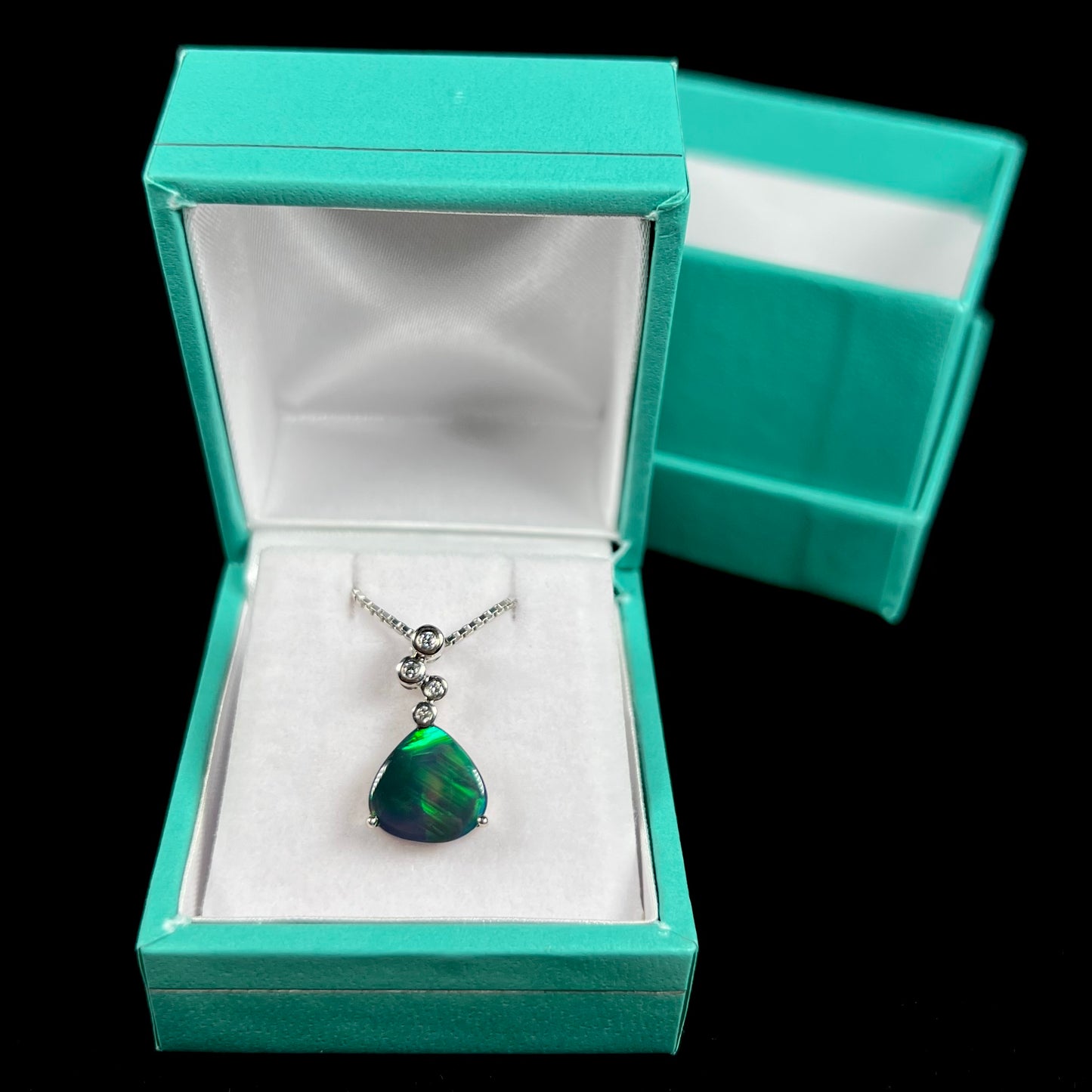 A black opal and diamond necklace displayed inside a blue pendant box against a black background, showing faint green color play.