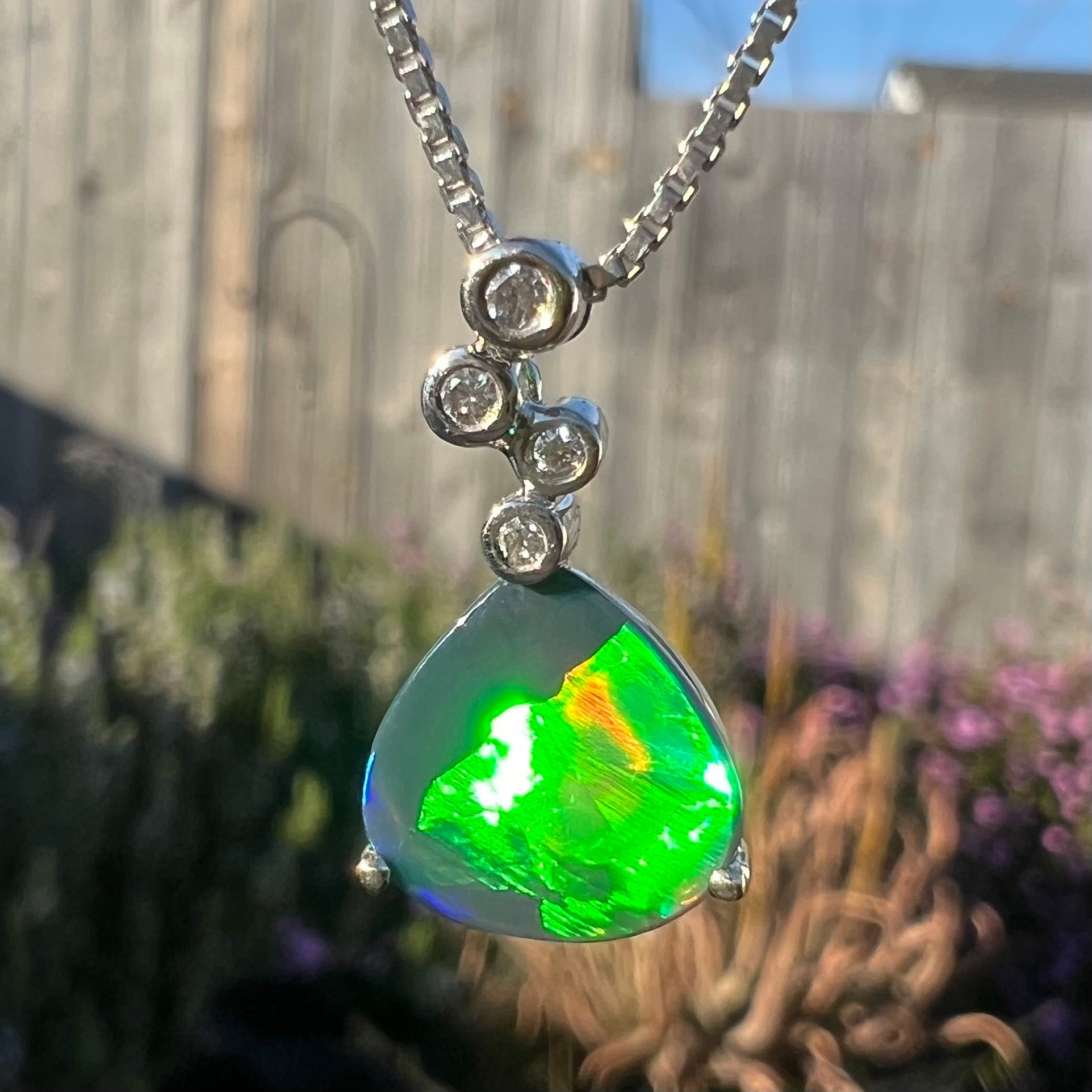 A macro close-up of a black opal and diamond necklace in full sunlight, revealing maximum green, red, and blue flashes.