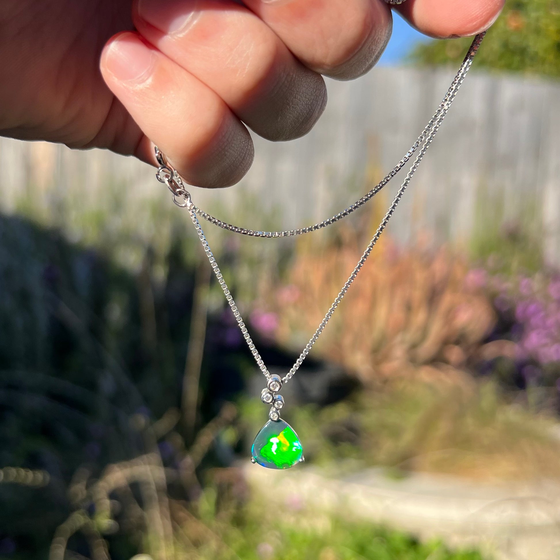 A black opal and diamond necklace dangling outdoors in sunlight, displaying a more vibrant green flash with a subtle red streak.