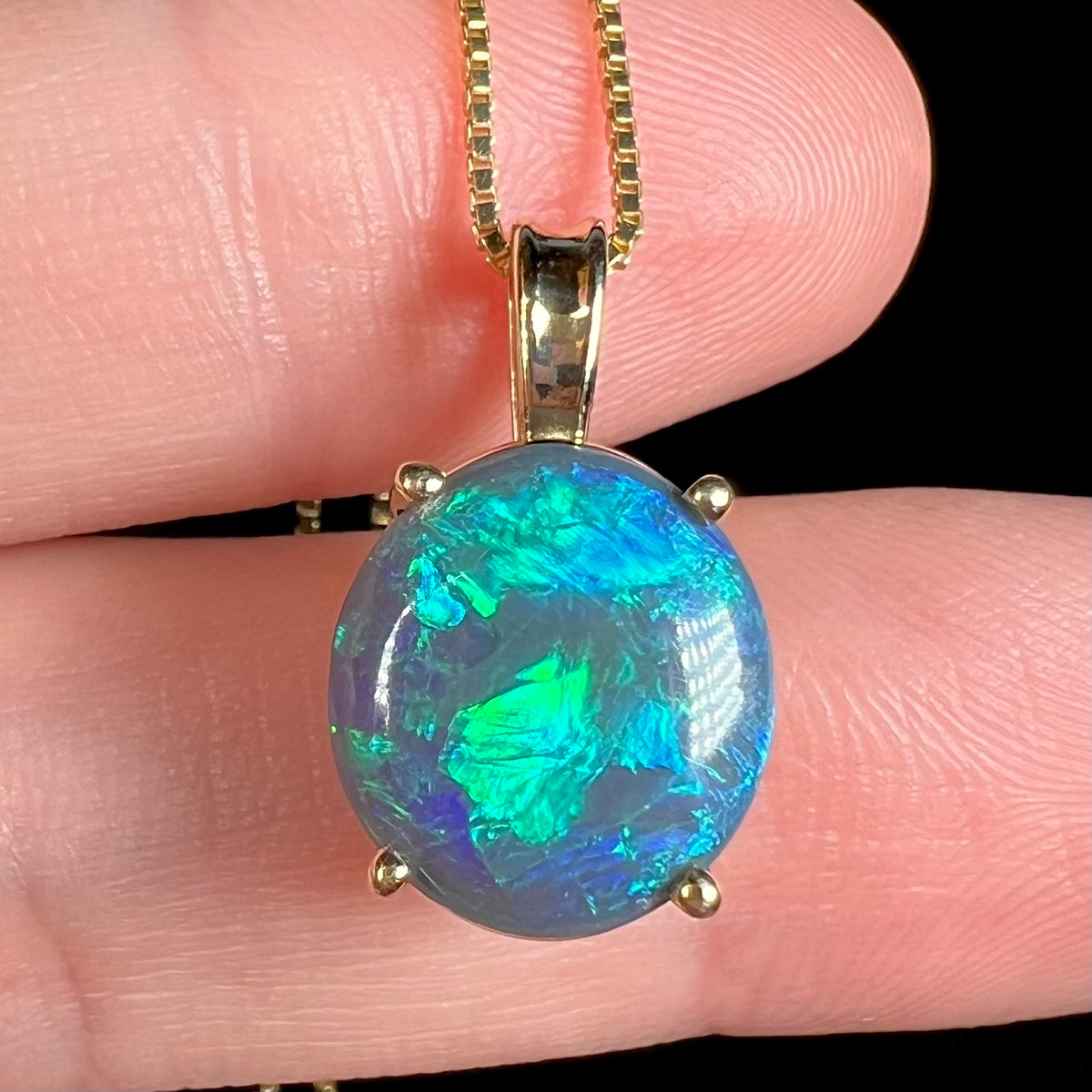 Close-up of a natural, solid Australian black opal pendant in 14k gold with green and blue flashes of color, resting on top of fingers