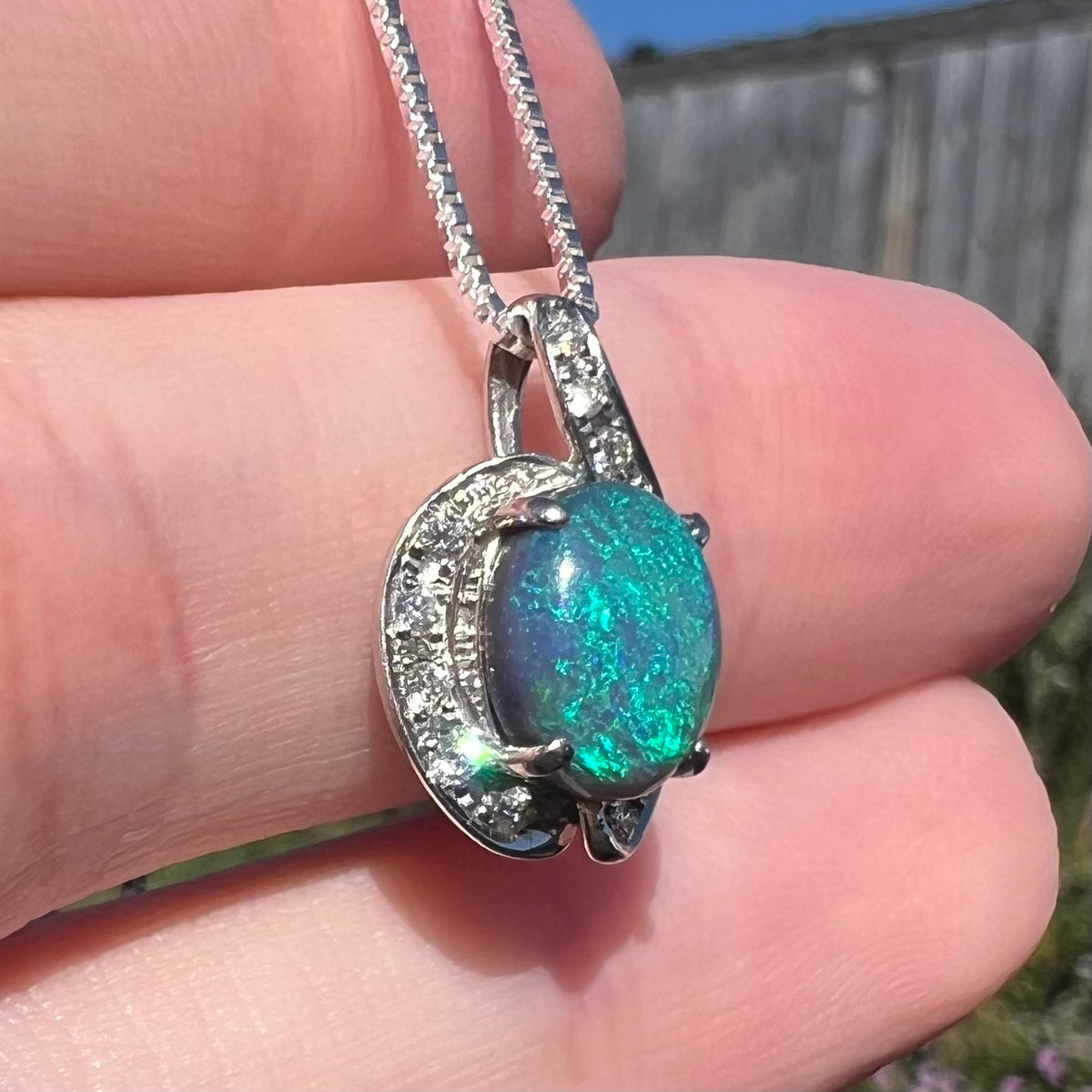 Blue and green flashes are seen in black opal pendant as a diamond accent sparkles, held in a hand under sunlight and viewed from an angle.